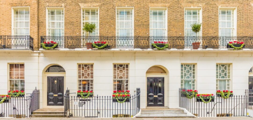 To refer to the 'Domicile' section of this tax guide for Hong Kong residents moving to the UK, a row of Kensington & Chelsea Edwardian style townhouses can be seen. It is a light and bright day, and there are red flowers on the gates and Juliette balconies. 