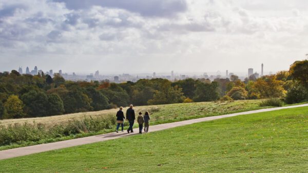 A family of four walking along a scenic path in a green park with panoramic views of the London skyline in the background. The image reflects the welcoming atmosphere for families joining skilled workers in the UK under the PBS Dependant visa scheme.